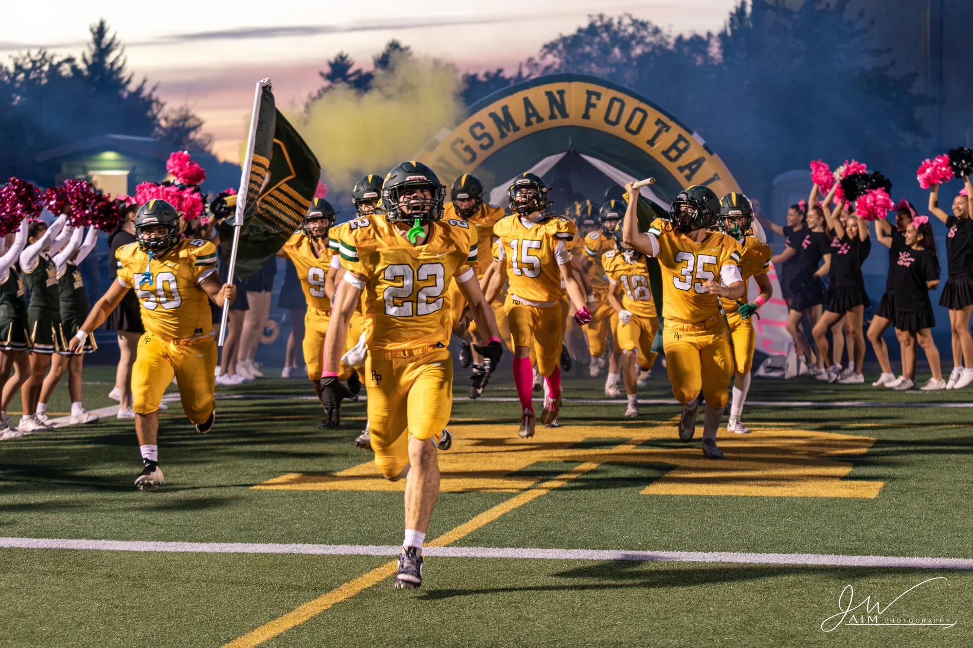 Kingsmen players in gold uniforms run onto the field through smoke at sunset